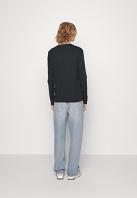 Sort langærmet skjorte parret med løstsiddende lyseblå jeans og sneakers, set bagfra mod en simpel hvid baggrund.