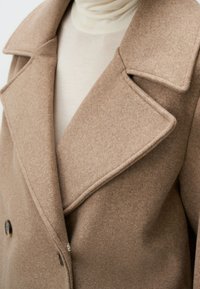 Cappotto in lana doppiopetto di colore marrone chiaro. Presenta ampi rever, tessuto testurizzato e due bottoni rotondi vicino all'orlo.
