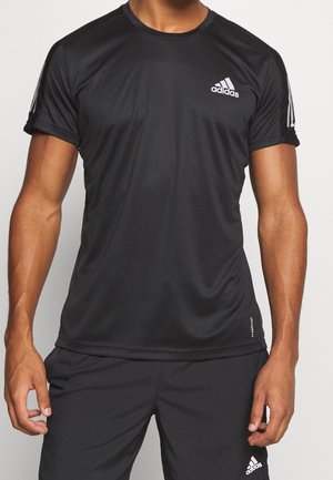 Mężczyzna ubrany w czarną sportową koszulkę Adidas z krótkim rękawem i czarne krótkie spodenki Adidas na jednolitym tle.