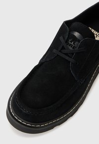 Scarpa in suede nera con design a lacci, punta tondeggiante e cuciture bianche a contrasto lungo la suola. Texture liscia e dettagli di branding minimi.