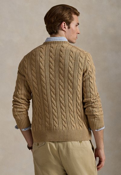 Man draagt een beige kabelgebreide trui over een blauw overhemd met kraag en beige broek, zijwaarts kijkend met handen in de zakken tegen een neutrale achtergrond.