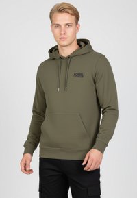 Giovane uomo che indossa un hoodie color verde oliva di Karl Lagerfeld con tasca frontale e pantaloni cargo neri, in piedi contro uno sfondo chiaro e semplice.