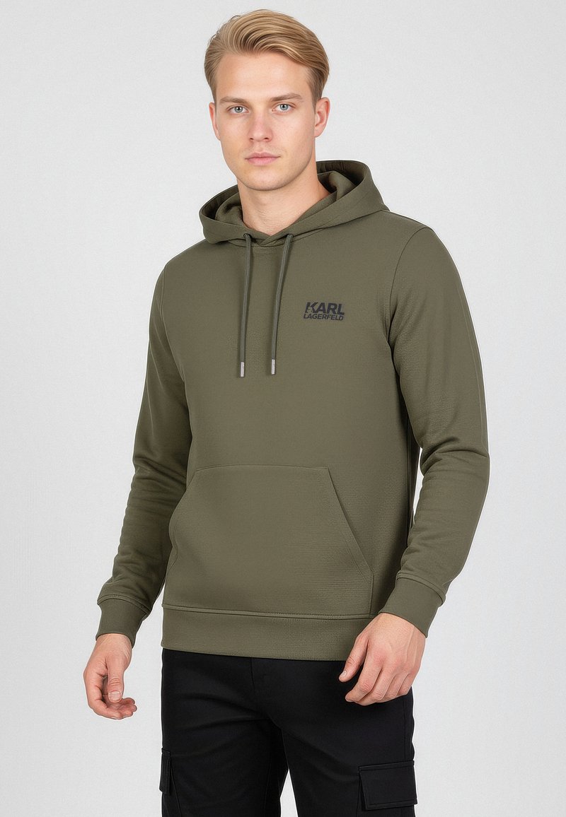 Giovane uomo che indossa un hoodie color verde oliva di Karl Lagerfeld con tasca frontale e pantaloni cargo neri, in piedi contro uno sfondo chiaro e semplice.