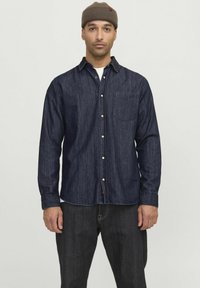 Camicia in denim blu scuro con design a bottoni, taschino singolo sul petto e maniche lunghe. Indossata con jeans scuri e un berretto marrone.