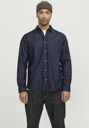 Donkerblauwe denim shirt met een knoopsluiting, een enkele borstzak en lange mouwen. Draagt het met donkerblauwe jeans en een bruine beanie.