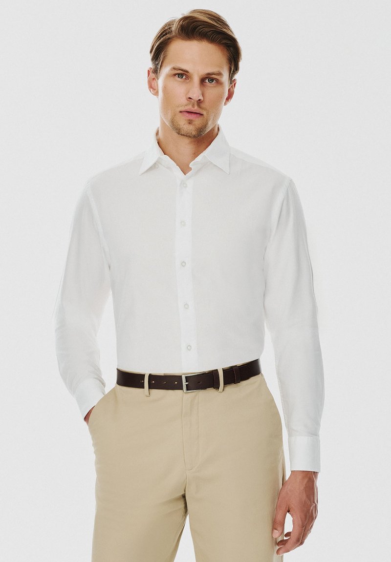 Biała koszula z długim rękawem, z wywijanym kołnierzykiem i zapięciem na guziki, zestawiona z beżowymi spodniami i brązowym skórzanym paskiem. Gładka tekstura materiału.