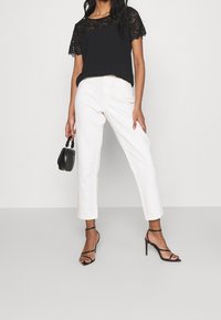 Haut en dentelle noir à manches courtes, associé à un pantalon blanc droit. Le mannequin tient un petit sac à main noir et porte des talons noirs à lanières.