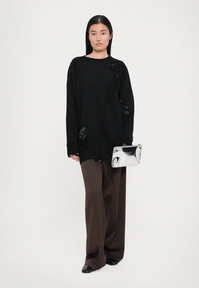 Black oversized sweater with textured patches, paired with brown wide-leg trousers and a shiny silver clutch. Solid color and relaxed fit.