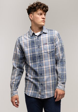 Młody mężczyzna w niebieskiej koszuli w kratkę, zapinanej na guziki, noszący białą koszulkę T-shirt, stojący na tle neutralnym, patrzący w bok.