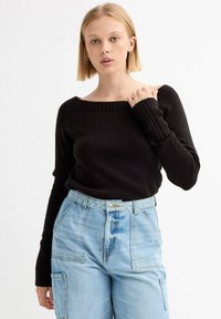 Suéter negro de punto acanalado con diseño de un solo hombro, combinado con jeans de mezclilla azul claro de cintura alta con bolsillos frontales.