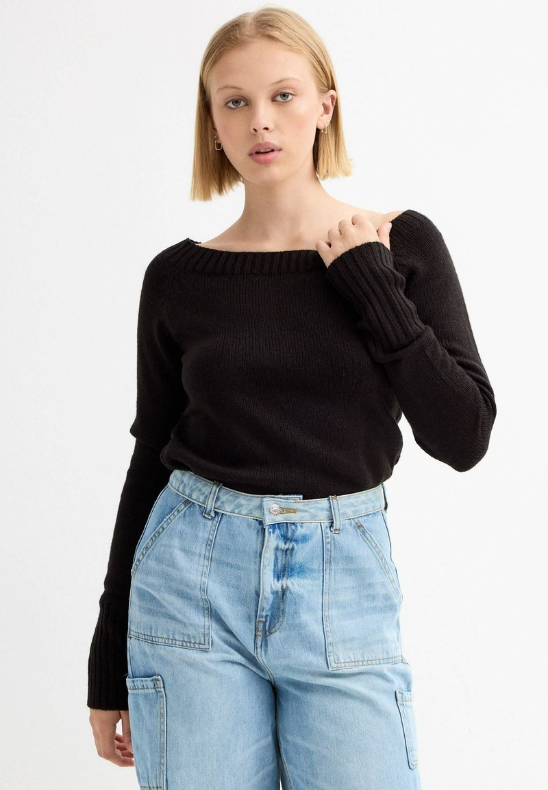 Suéter negro de punto acanalado con diseño de un solo hombro, combinado con jeans de mezclilla azul claro de cintura alta con bolsillos frontales.