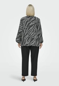 Grå blus med zebramönster, långa ärmar och avslappnad passform, tillsammans med svarta byxor och klacksandaler. Bakifrån visad.