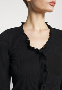 Svart ribbad cardigan med en volangkrage, långa ärmar och en figurnära silhuett. Texturen är mjuk och elastisk.