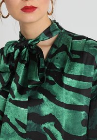 Green and black patterned blouse with a tie neckline, featuring a smooth fabric texture and bold, abstract stripes.