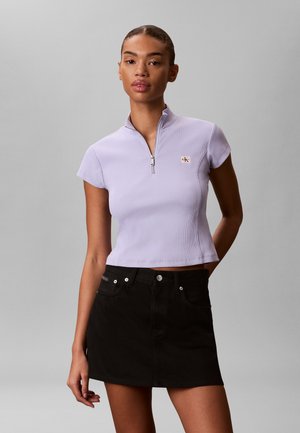 Kobieta ubrana w jasnofioletowy krótki top z zamkiem i czarną mini spódniczkę, stojąca z jedną ręką za plecami na jednolitym tle.