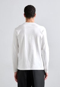 Hvid langærmet t-shirt lavet af bomuld, med rund hals og et lille rektangulærtmærke på bagsiden, parret med mørke bukser.
