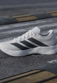 Chaussure de sport blanche avec des bandes noires placée sur une surface asphaltée texturée près d’un passage piéton et de ralentisseurs.