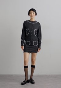 Rochie tricotată neagră cu desene albe pe buzunare, mâneci lungi, front cu butoane, formă ajustată, asortată cu ciorapi până la genunchi și pantofi negri.