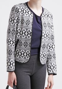 Černobílá vzorovaná bunda s geometrickými designy, žebrovanou texturou a límcem ve vojenském stylu, v kombinaci s námořnickým topem a šedými kalhotami.