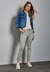 Denim jack in blauw, lichtgrijze broek en witte sneakers. Model staat casual met opgerolde beenopeningen. Eenvoudig T-shirt eronder.