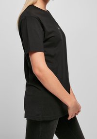 Černé bavlněné tričko s kulatým výstřihem a krátkými rukávy, který má malou grafiku na hrudi. Boční pohled ukazuje uvolněný střih.