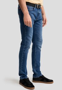 Slim-fit blue jeans made of denim, featuring five pockets, stitched accents, and a black belt. Worn with black sneakers.