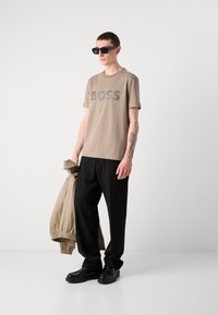 Ruda medvilninė marškinėliai su "BOSS" logotipu, derinami su juodomis kelnėmis, šviesiai ruda striuke ir juodais slip-on batais; dėvi tamsius akinius nuo saulės.