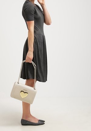 Nainen pukeutuneena tummanharmaaseen lyhythihaiseen mekkoon, kantaa vaaleanpunaista käsilaukkua, jossa on kultainen sydänkoriste, seisoo neutraalilla vaalealla taustalla.