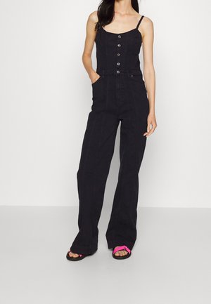 Frau trägt schwarzen, knöpfenlosen, ärmellosen Jumpsuit mit weitem Bein und rosa Sandalen, stehend vor einem schlichten weißen Hintergrund.