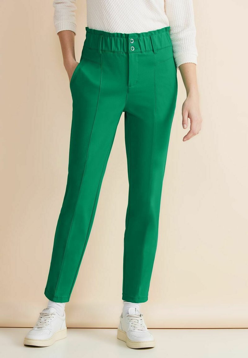 Zelené kalhoty na míru s vysokým elastickým pasem, dvěma knoflíky, bočními kapsami a slim fit. Stylizováno s bílými teniskami.