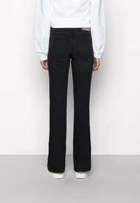 Persona con pantalones vaqueros negros de cintura alta y zapatillas blancas, de pie contra un fondo claro y liso, vista desde atrás.