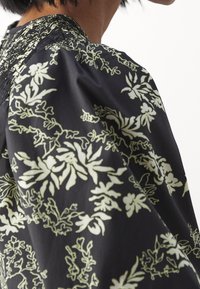 Gros plan d'une personne portant un vêtement noir avec des motifs floraux crème et un détail froncé près de l'encolure.