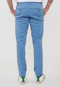 Pantaloni in cotone blu con vestibilità slim, dotati di due patte sui taschini posteriori e una superficie liscia. Indossati con sneakers bianche e verdi.