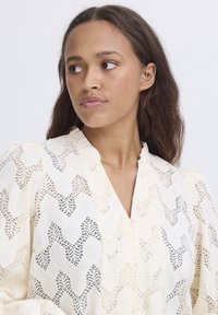 Lace blouse in cream, featuring a geometric pattern with subtle black accents. It has a v-neck and button closure with a textured finish.