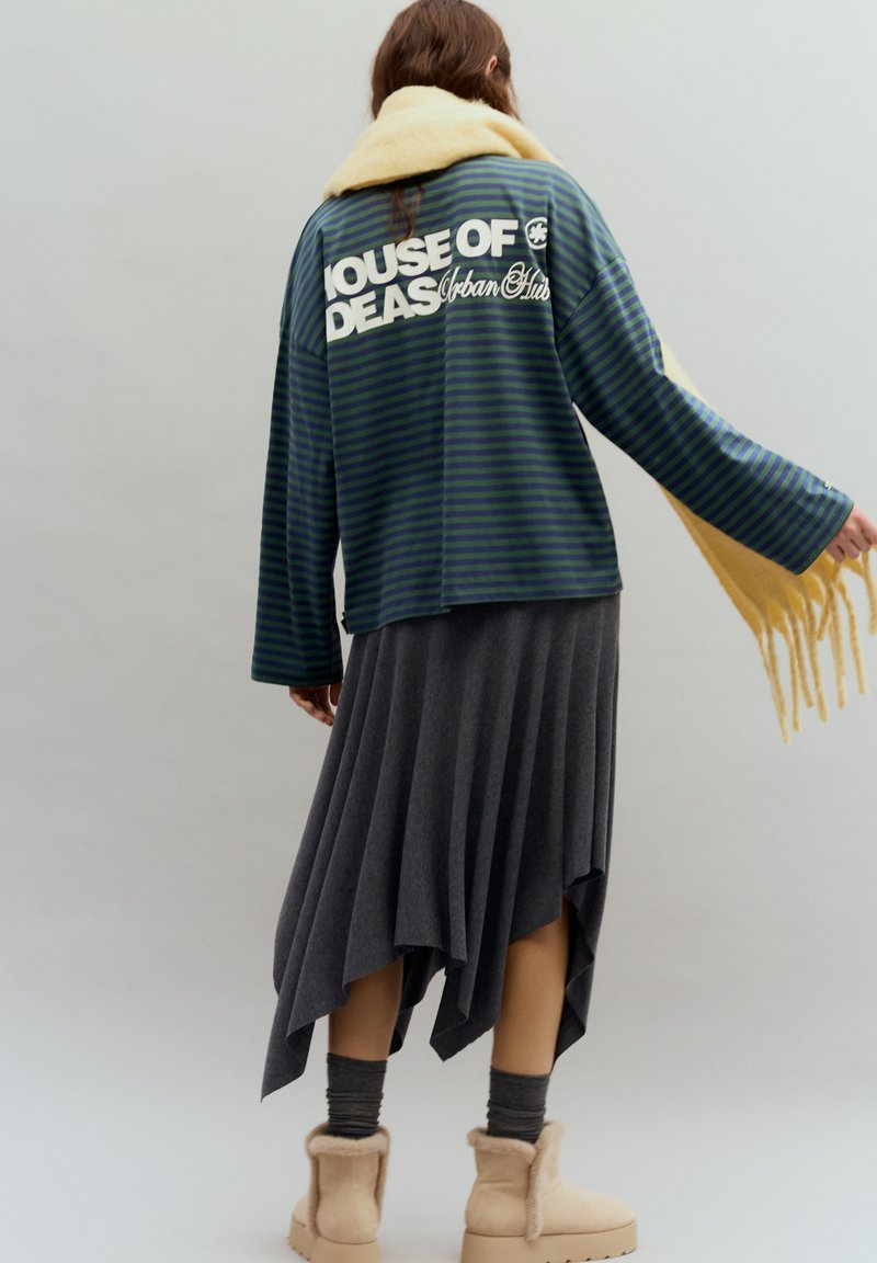 Zöld és tengerészkék csíkos pulóver "HOUSE OF IDEAS" felirattal, szürke aszimmetrikus szoknyával és bézs színű, szőrmével bélelt csizmával párosítva, sárga sállal átdobva.