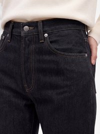 Nahaufnahme einer Person, die schwarze Denim-Jeans mit sichtbaren Nähten, einem vorderen Knopf, Gürtelschlaufen und einem Teil eines hellfarbenen Pullovers trägt.