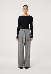 Vrouw die rechtop staat en een zwarte ribgebreide longsleeve top en hoge, wijde grijs gestreepte broek met een strikceintuur draagt.