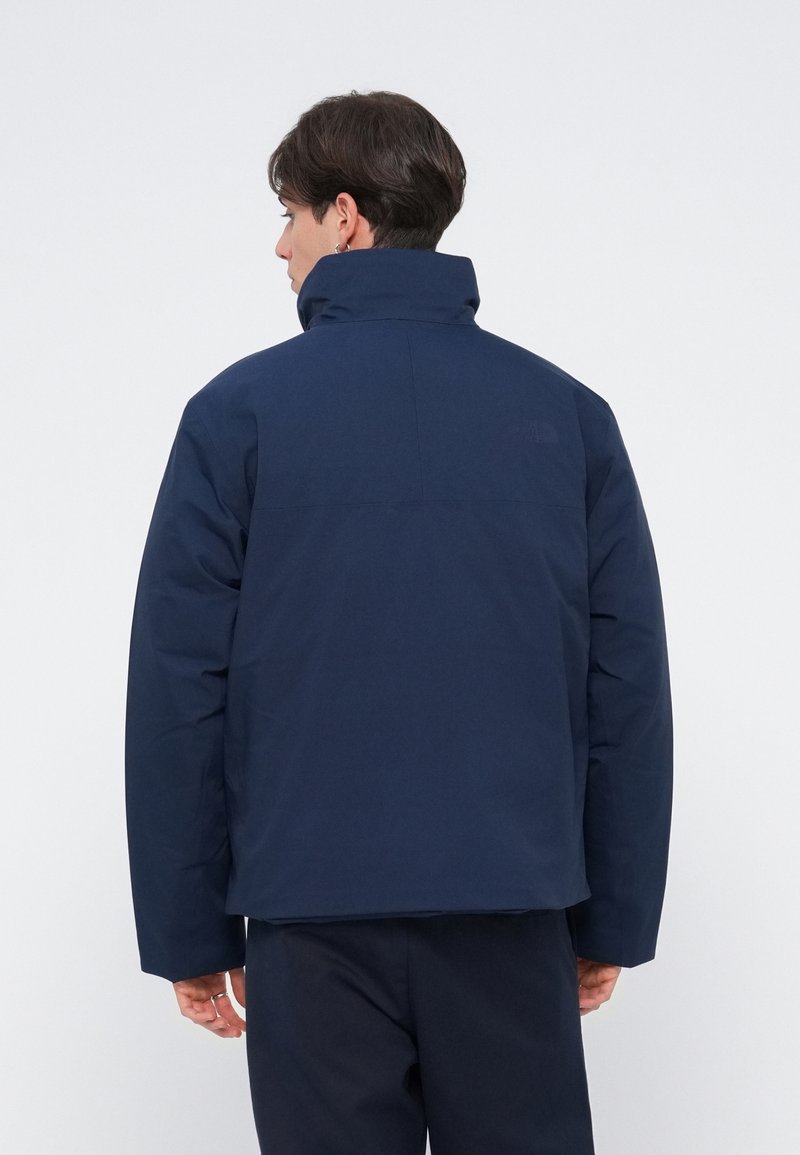 Doudoune bleu marine avec un col haut, présentant un design épuré et des coutures minimalistes. Texture lisse, tissu durable, sans quincaillerie visible.