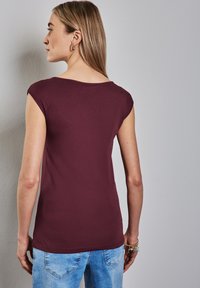 Burgundské přiléhavé tričko s krátkými rukávy, kulatým výstřihem a hladkým materiálem. Modelka má na sobě modré džíny, které předvádějí zadní pohled na tričko.