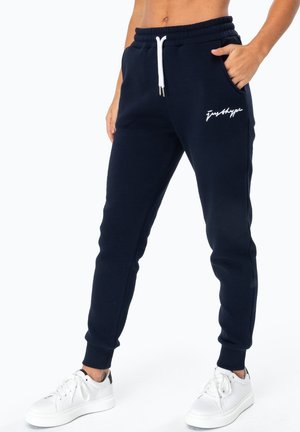 Marineblauwe joggingbroek met witte trekkoord en "justhype" logo op linker dij, gedragen met witte sneakers en hand in zak.