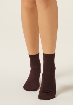 Braune Baumwoll-Knöchelsocken, weich strukturiert mit einem gerippten Bündchen, getragen an bloßen Beinen vor einem hellen Hintergrund.