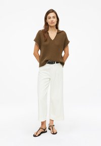 Mujer con un top marrón de punto de manga corta, pantalones blancos de pierna ancha, cinturón negro y sandalias negras, de pie con las manos detrás de la espalda.