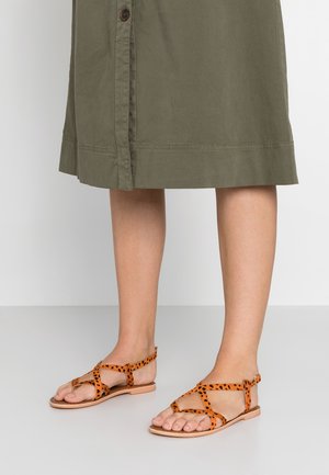 Piernas con falda verde oliva y sandalias planas de punta abierta con tiras salpicadas de naranja y negro sobre un suelo blanco.