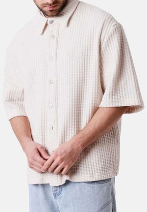 Homme portant une chemise à manches courtes texturée de couleur crème et un jean bleu clair, en train de boutonner les boutons du bas.