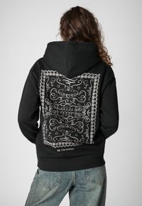 Zwarte hoodie met een groot wit, ingewikkeld patroon op de achterkant en de tekst "BE THE ENERGY" vlak bij de zoom.