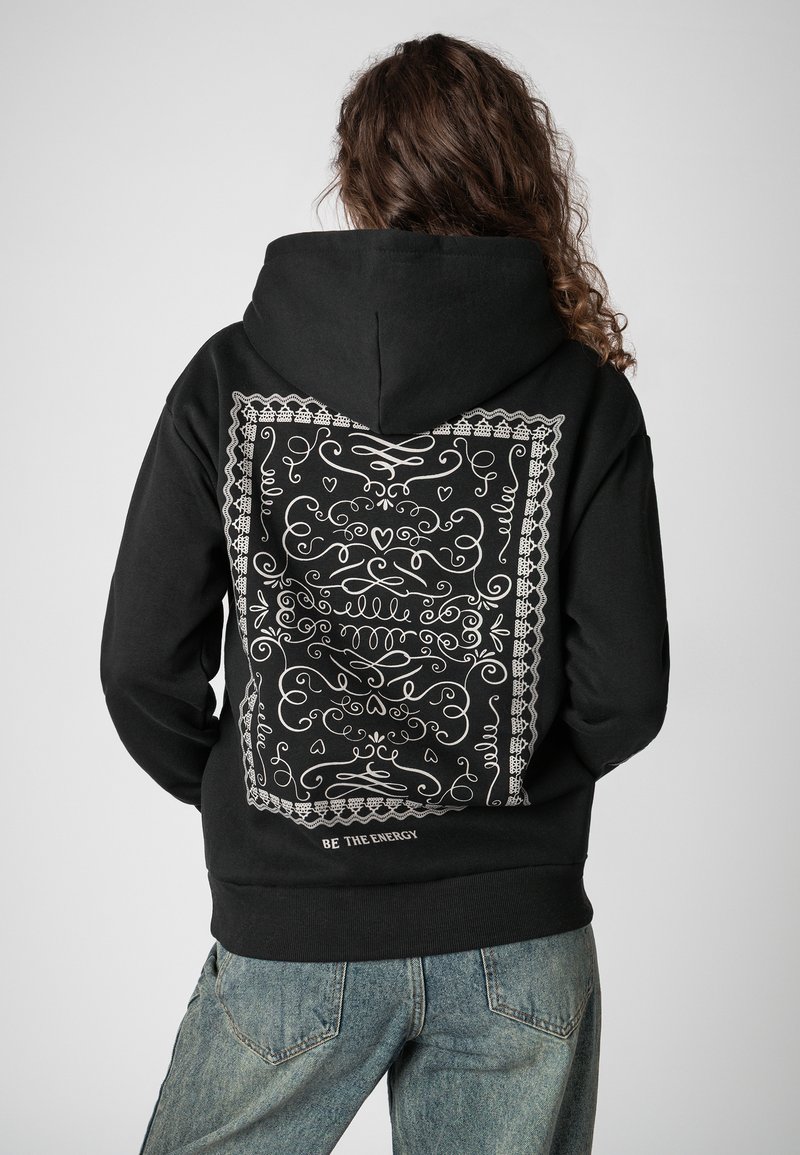 Zwarte hoodie met een groot wit, ingewikkeld patroon op de achterkant en de tekst "BE THE ENERGY" vlak bij de zoom.
