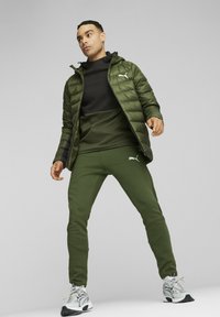 Grøn isoleret jakke, sort top, grønne bukser og hvide sneakers. Har logo detaljer på jakken og bukserne. Glat tekstur.