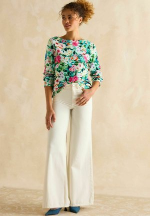 Bluson mit langen Ärmeln im Floral-Design auf grünem Hintergrund, verziert mit rosa, weißen und blauen Blumen; kombiniert mit hoch taillierten, weiten weißen Hosen.