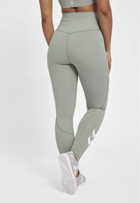 Höga gröna leggings tillverkade av elastiskt tyg, med sömmar och vita logotypdetaljer vid anklarna, som är ihop med ljusa sneakers.