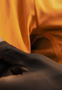 Close-up of a person wearing a black glove touching the orange fabric of a garment with detailed stitching and mesh lining.